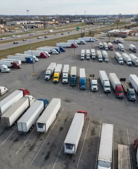 truck stop parking area near a major highway. Multiple semi-trucks with trailers are parked in marked parking spaces