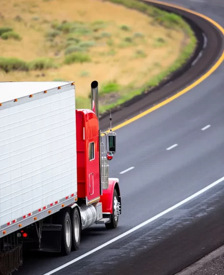 Rear three-quarter view of a red semi-truck with a white box trailer driving along a curving highway, bordered by grassy hills and open landscape, conveying long-haul freight transport.