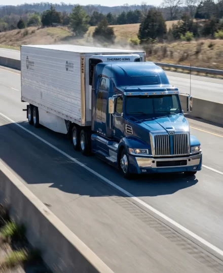Western Star semi-truck driving on an American highway, hauling a long refrigerated trailer. The truck is captured in motion, traveling at highway speed.