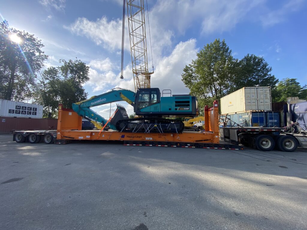 Tracked excavator secured on an orange heavy-haul trailer, positioned beneath a crane boom in a container yard with shipping containers, trees, and clear blue sky.