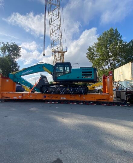 Tracked excavator secured on an orange heavy-haul trailer, positioned beneath a crane boom in a container yard with shipping containers, trees, and clear blue sky.