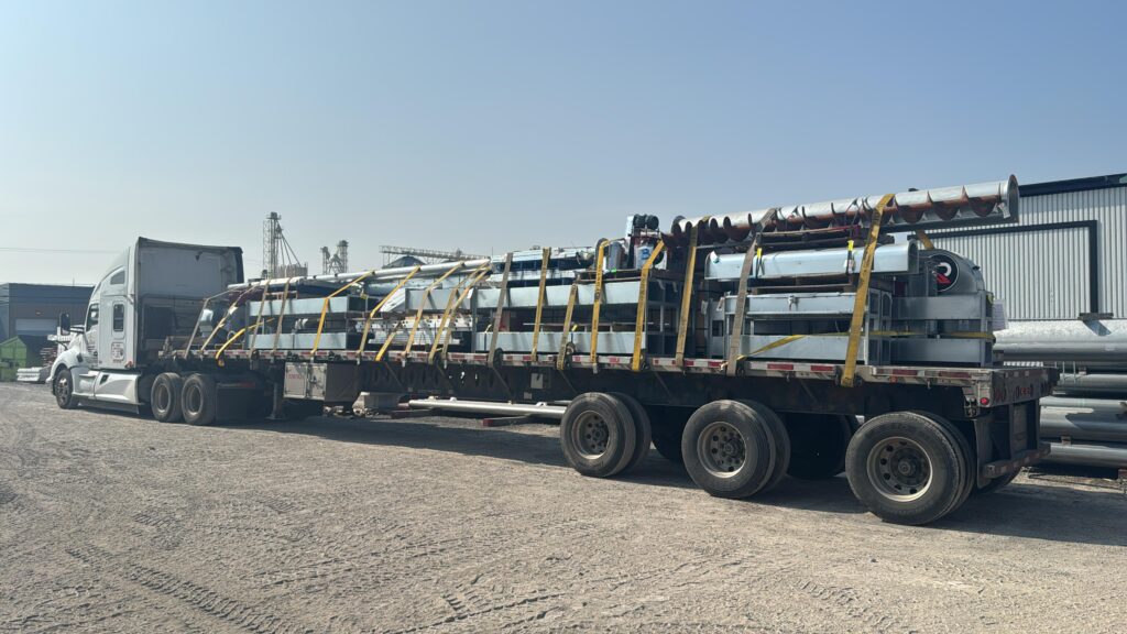 Flatbed semi truck parked in an industrial yard, hauling long metal augers and equipment secured with yellow straps, with warehouses and machinery in the background on a clear day.