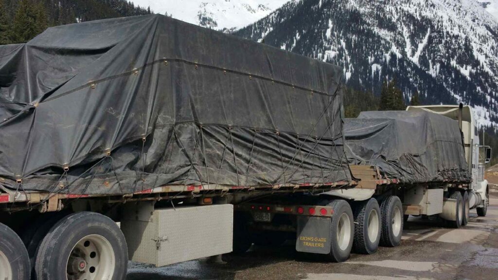 Two flatbed trucks parked outdoors, each carrying oversized cargo fully covered with dark tarps and secured with straps. The trucks are positioned on a roadside with snow-covered mountains and evergreen trees in the background, suggesting long-distance transport through a mountainous region.