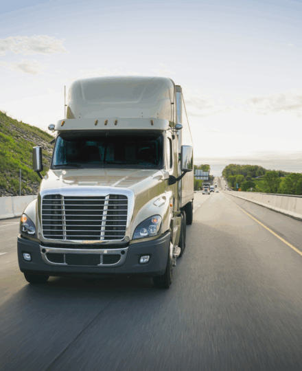 Front view of a white aerodynamic semi-truck driving on a multi-lane highway, with chrome grille and side mirrors visible, rolling through a semi-rural landscape with grassy embankments and trees under a bright, lightly cloudy sky.