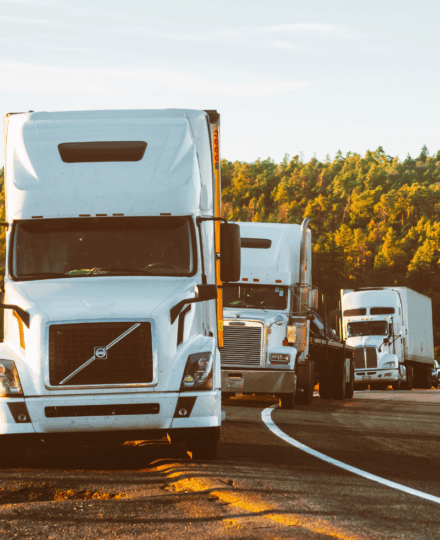 Front view of a white aerodynamic semi-truck driving on a multi-lane highway, with chrome grille and side mirrors visible, rolling through a semi-rural landscape with grassy embankments and trees under a bright, lightly cloudy sky.