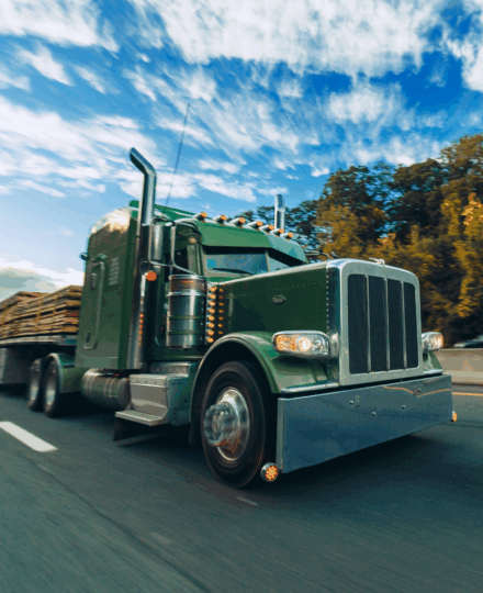 Green long-nose Peterbilt semi truck with a classic and chrome exhaust stacks driving on a highway, hauling a flatbed load with motion blur, autumn trees lining the road under a dramatic blue sky.