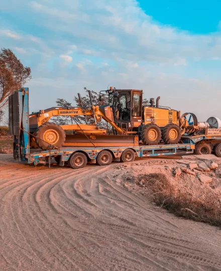 Yellow motor grader secured on a multi-axle flatbed trailer, parked on a dirt construction site with tire tracks, sparse trees, and a partly cloudy sky at golden hour.
