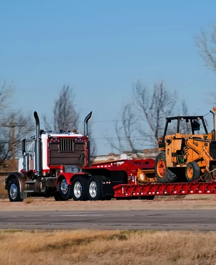 Red heavy-duty semi truck hauling a yellow construction loader on a red lowboy trailer, traveling along a rural roadway with bare trees and open fields under a clear blue sky.