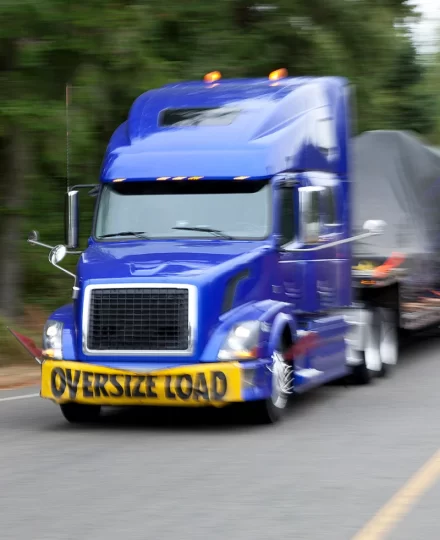 Blue semi-truck with an OVERSIZE LOAD banner mounted on the front, hauling a covered load on a flatbed trailer while driving on a two-lane road bordered by trees, with motion blur indicating movement.